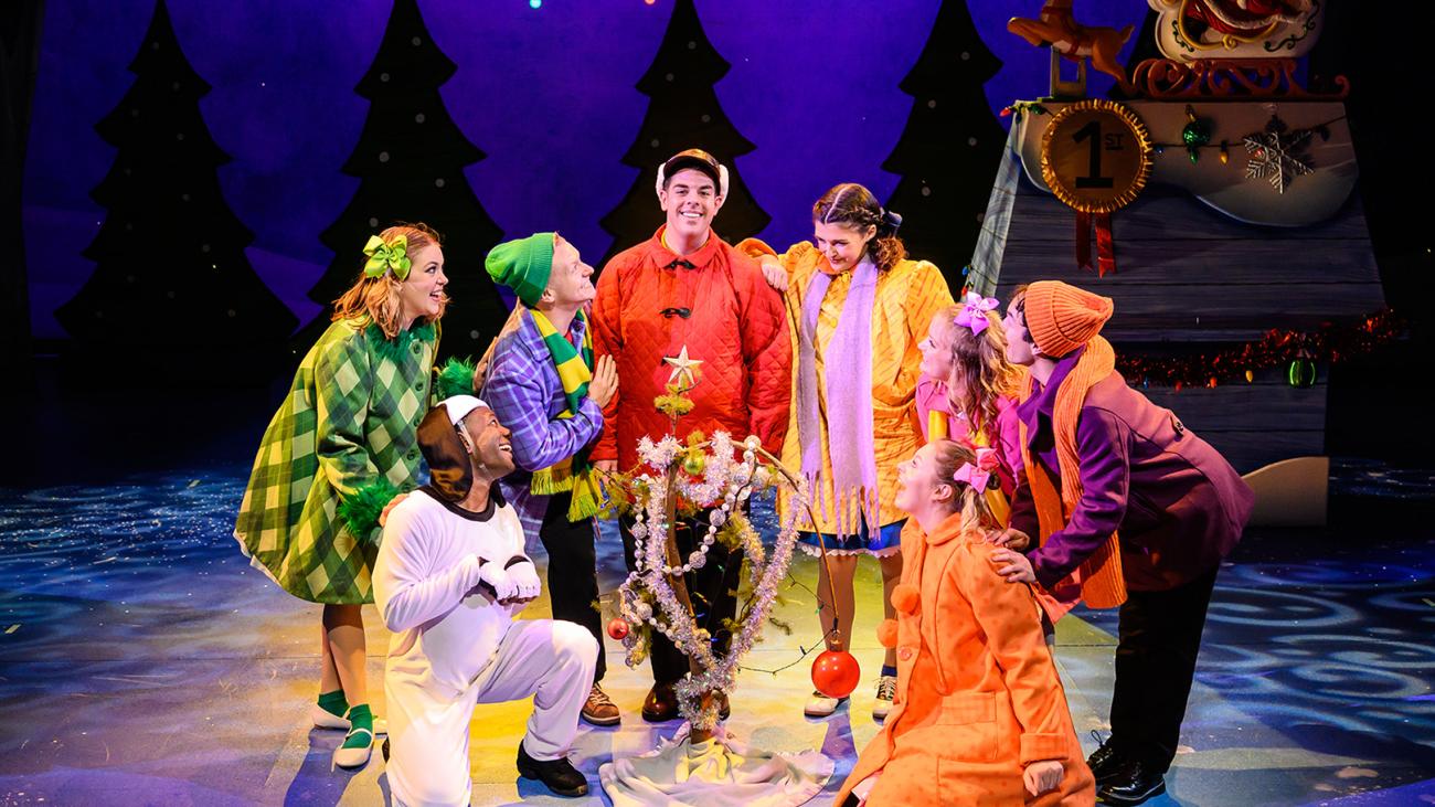 A group of actors dressed as the charaters from Charlie Brown gather in a circle around a small branch decorated as a christmas tree.